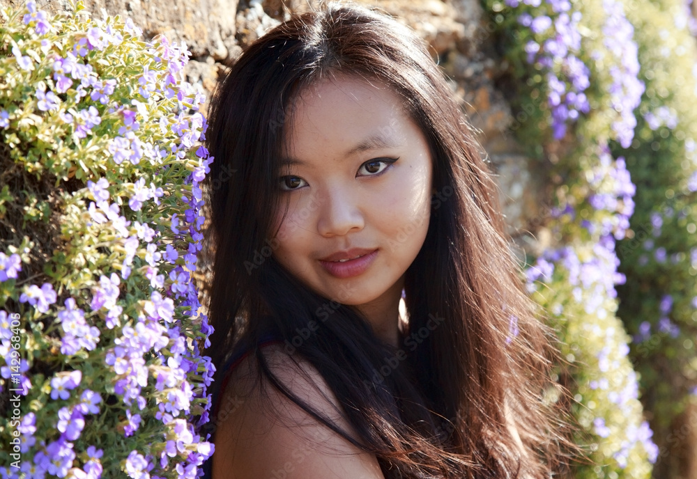 Naklejka premium Beautiful potrait of a Chinese, Oriental young woman with long dark hair sitting against a wall of purple flowers