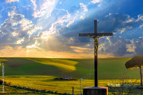 Jesus Christ on the cross by the road in the fields at sunset, with drammatic clouds and sun rays on the sky. photo