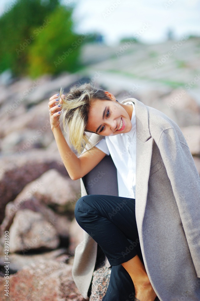 Portrait of young beautiful smiling girl sitting on rock, on coast to coat draped over shoulders, on blurred background, close up.