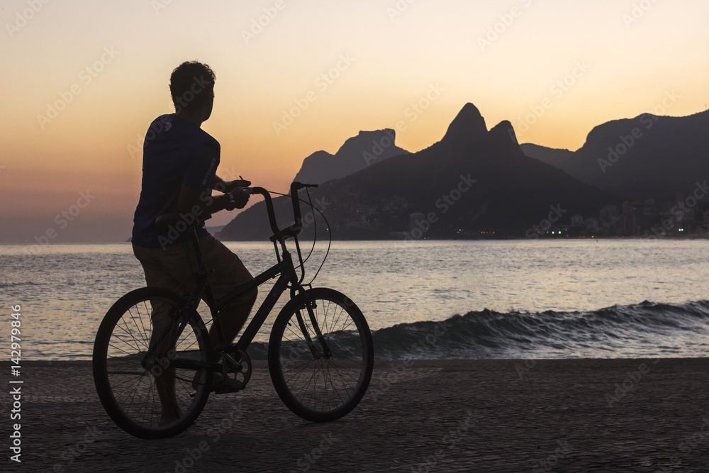 Fototapeta premium Man on a bicycle watching the sunset behind Dois Irmãos rock on Ipanema beach