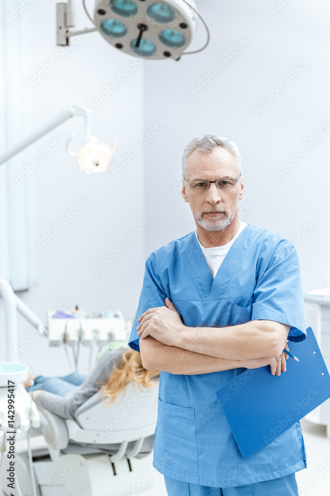 senior dentist in uniform with clipboard in dental clinic with patient behind senior dentist in uniform with clipboard in dental clinic with patient behind