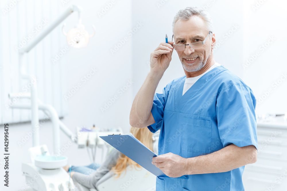 senior dentist in uniform with clipboard in dental clinic with patient behind