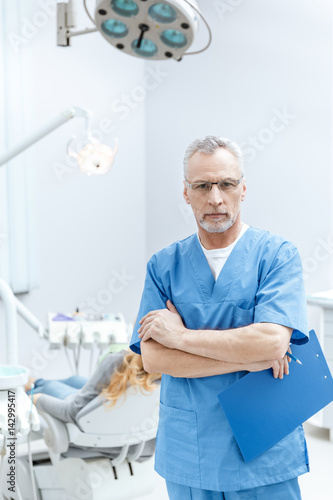 senior dentist in uniform with clipboard in dental clinic with patient behind