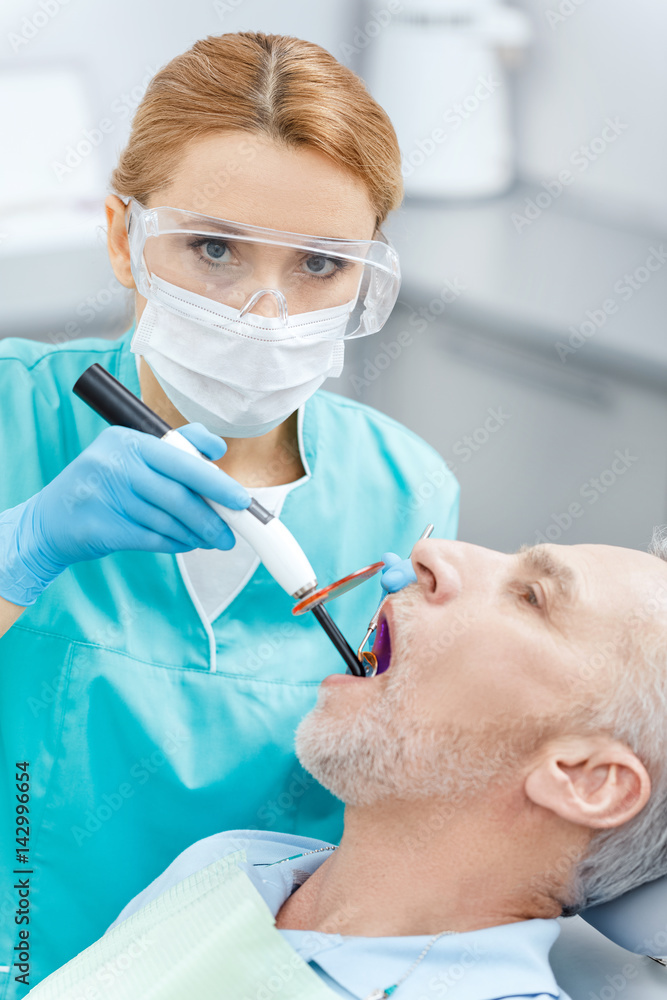 Professional dentist using dental curing UV lamp on teeth of mature patient Professional dentist using dental curing UV lamp on teeth of mature patient
