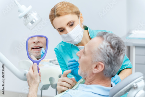 Mature patient looking at mirror while professional dentist checking teeth