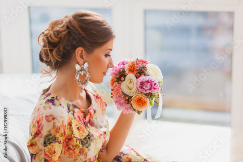 Beautiful girl in a long dress sitting near the sofa and sniffing bouquet