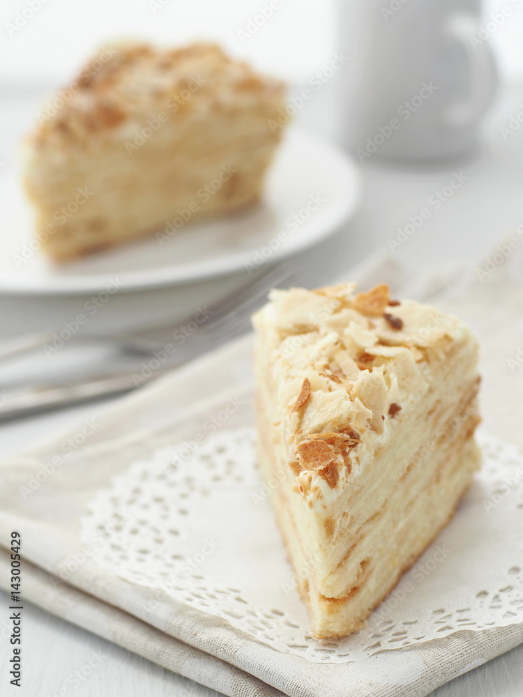 Piece of multi layered cake close-up. Mille feuille dessert. Crumbs decorated torte on white doily upon wooden table. Food background, blur. Selective focus on the front. Piece of multi layered cake close-up. Mille feuille dessert. Crumbs decorated torte on white doily upon wooden table. Food background, blur. Selective focus on the front.