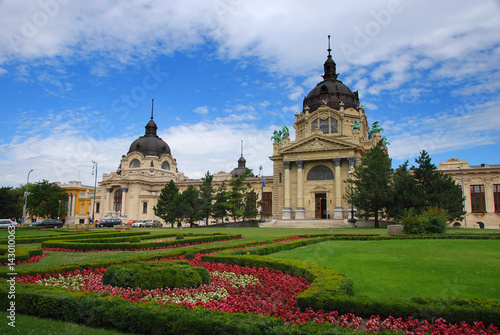 A large church with two towers. A beautiful and wide garden with flowers is in front of it. The day is sunny.