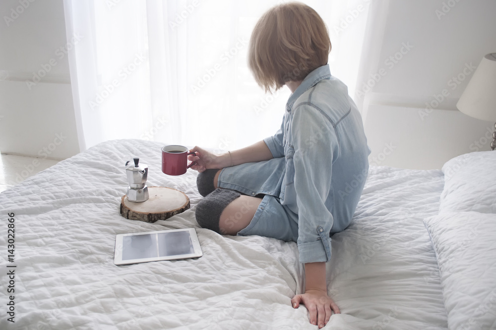 Girl with cup in her hand, kettle and tablet on a white bed looking in window Girl with cup in her hand, kettle and tablet on a white bed looking in window