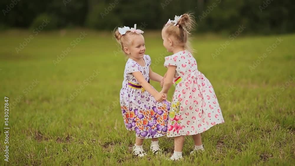 Two cute sisters twins frolicking on a green meadow in summer day. Holding hands smile at each other. Two cute sisters twins frolicking on a green meadow in summer day. Holding hands smile at each other.