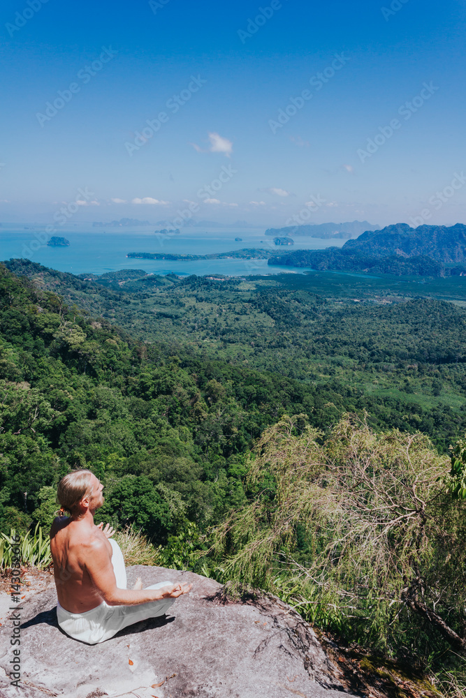 Young male practicing yoga on nature. From above shot of man in white practicing yoga pose on rock against beautiful tropical views.
