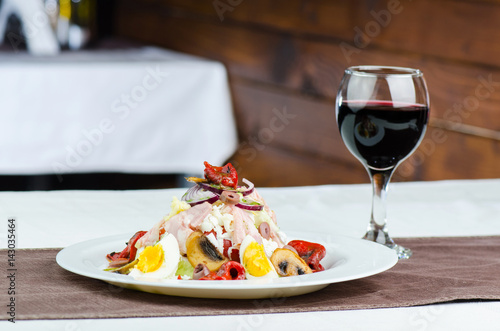 Fresh Salad With Mushrooms, Grilled Vegetables, Boiled Eggs, White Cheese, Olive And Ham With Glass Of Red Wine On Wooden Background