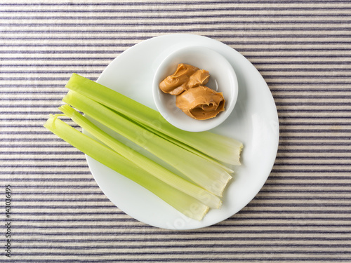 Top view of celery stalks with peanut butter in a small bowl on a white plate atop a blue striped tablecloth.
