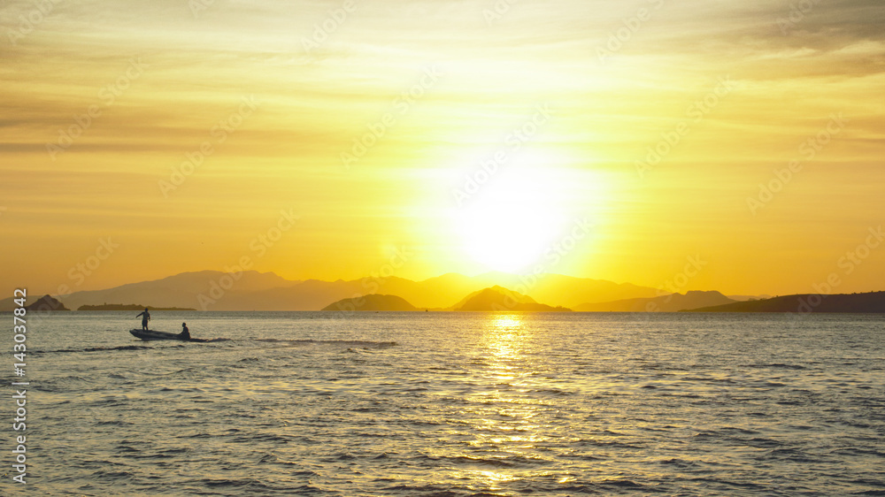 Silhouette of two fishermen on a small fishing boat with outboard motor on the ocean at nightfall with an orange sunset at the background in asia.