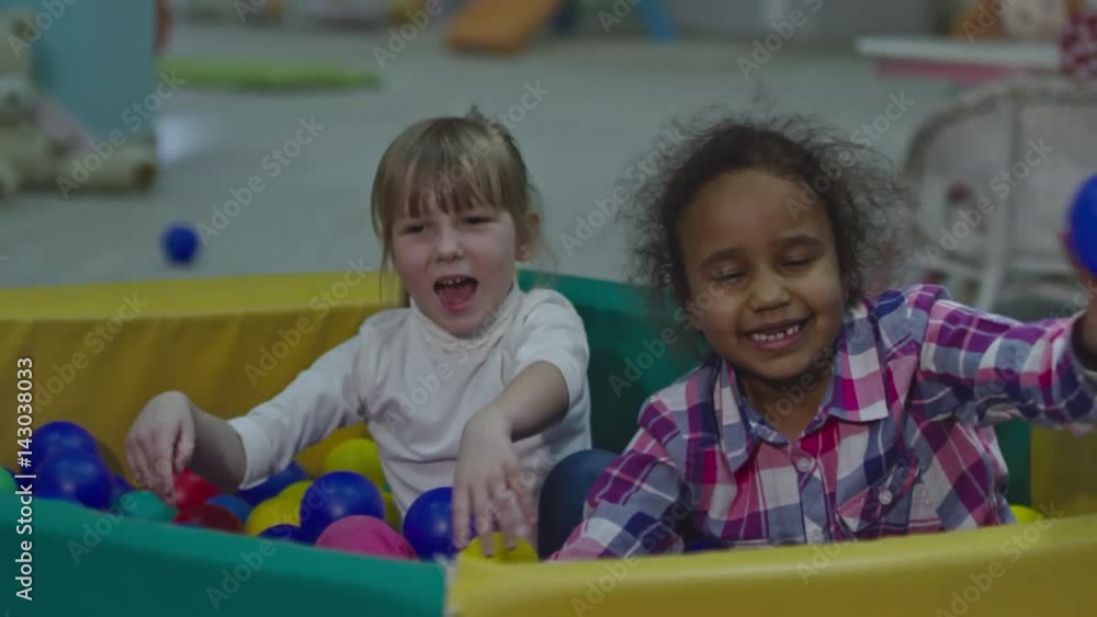 Two little African and Caucasian girls sitting in colorful ball pool in children play room and throwing balls in slow motion Two little African and Caucasian girls sitting in colorful ball pool in children play room and throwing balls in slow motion