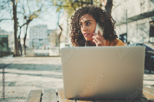 Serious woman calling with cell telephone while sitting in street cafe, attractive brunette curly lady having talking on smart phone while work on her modern laptop with copy space for your text