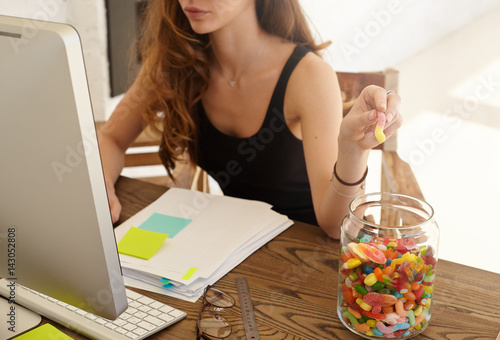 Cropped shot of the Caucasian female office worker taking marmalade from the big jar at work. The girl eats candies for improvement of work of a brain during hard work over the financial statement