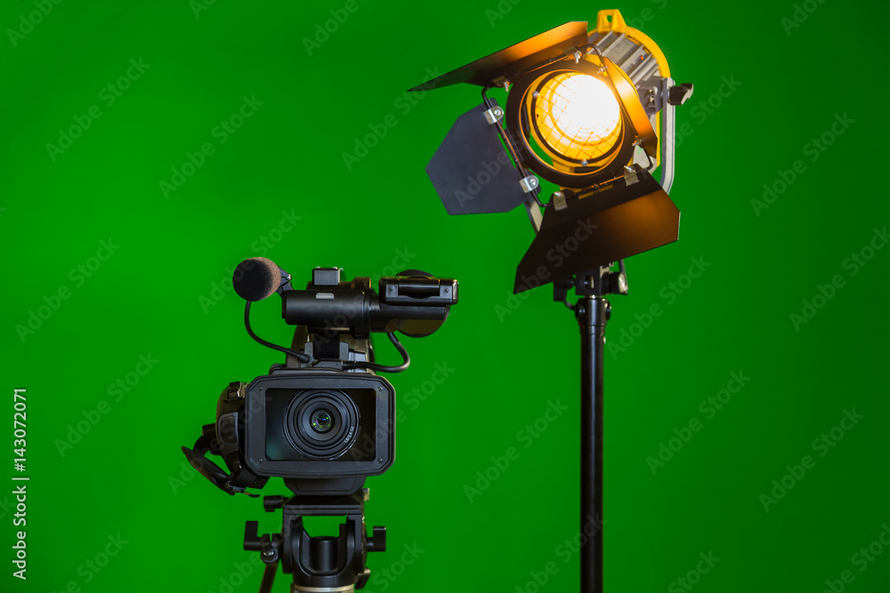 A video camera and a spotlight with a Fresnel lens on a green background. Filming in the interior. The chroma key.