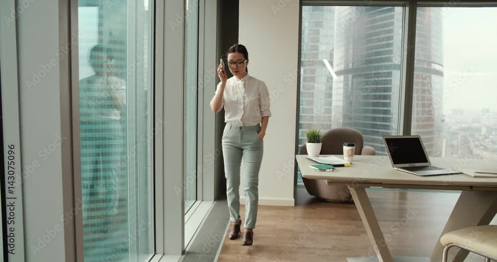 Asian girl busy talking on the phone walks in a modern office, a beautiful view of the skyscrapers outside the window Asian girl busy talking on the phone walks in a modern office, a beautiful view of the skyscrapers outside the window