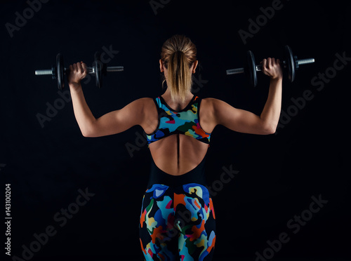 Young female bodybuilder with perfect strong muscular body wearing sportswear tracksuit exercising with weights. Rearview.