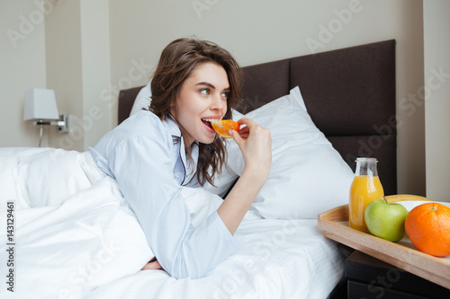 Happy young lady under blanket eating orange.