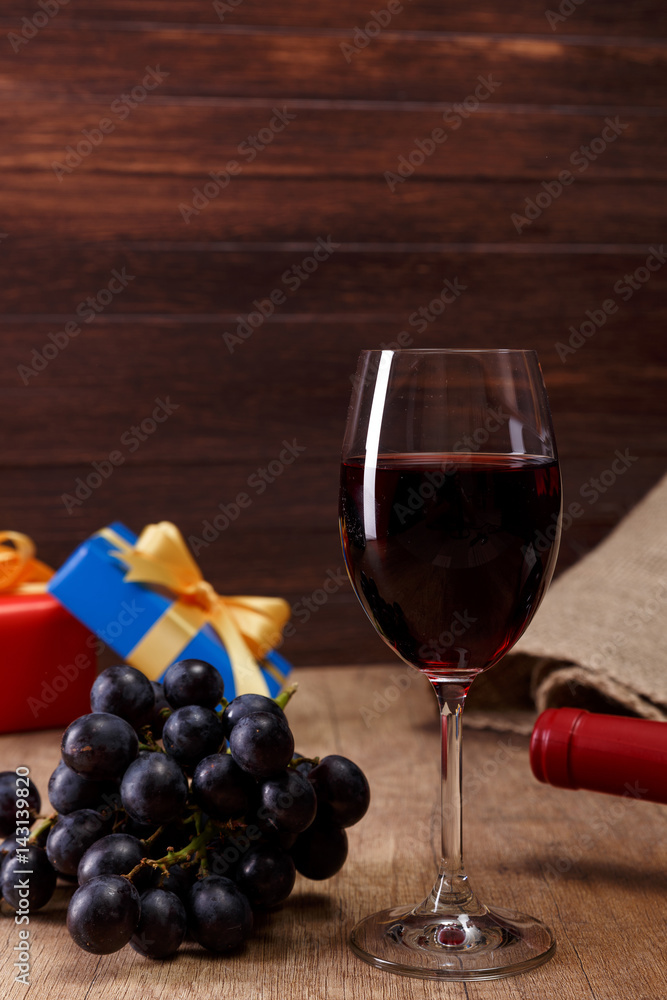 Red wine bottle. Wineglass with dark grapes branch. Gift box with bow on rustic wooden background.