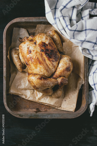 Grilled baked whole organic chicken on backing paper in old oven tray with white kitchen towel over black burnt wooden background. Top view with space.