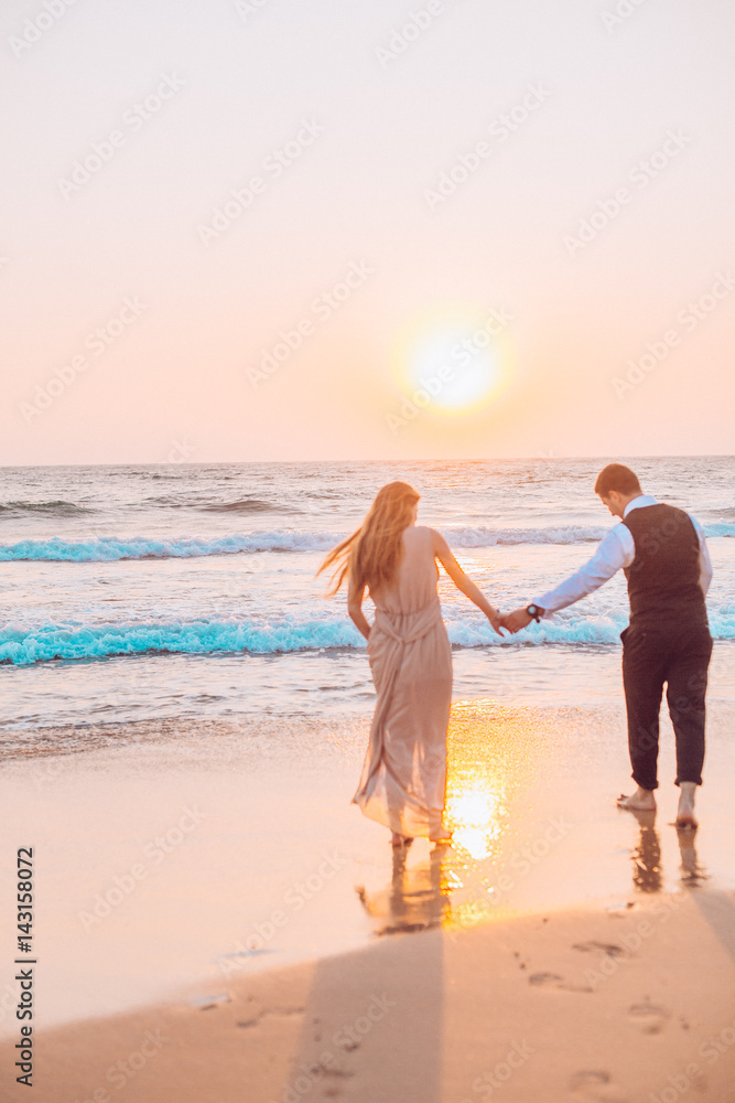 Honeymoon couple romantic in love at beach sunset. Newlywed happy young couple holding hands enjoying ocean sunset during travel holidays vacation getaway.