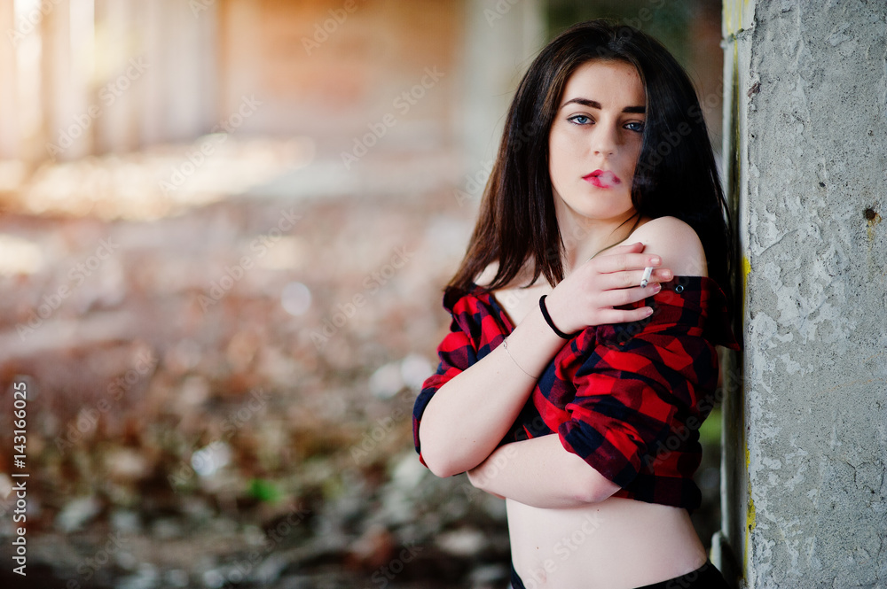 Obraz premium Portrait smoking girl with red lips wearing a red checkered shirt with bare shoulders posed sexy background abadoned place.