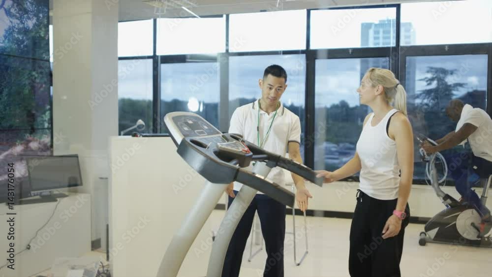 Woman using treadmill in modern gym with view of natural environment on video screen. Personal trainer monitors her progress. Woman using treadmill in modern gym with view of natural environment on video screen. Personal trainer monitors her progress.