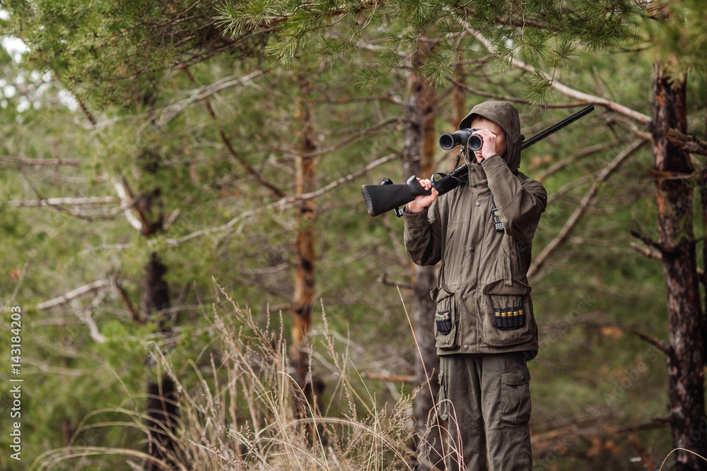 Fototapeta premium male hunter with binoculars ready to hunt, holding gun and walking in forest.