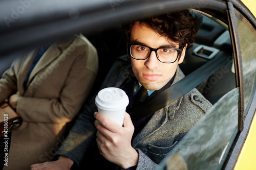 Portrait of confident Middle-Eastern businessman riding in backseat of car smiling cheerfully looking at camera out of window lit by sunlight, holding coffee cup