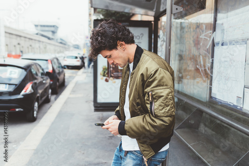Handsome young man at bus stop using smart phone hand hold - social network, commuting, technology concept