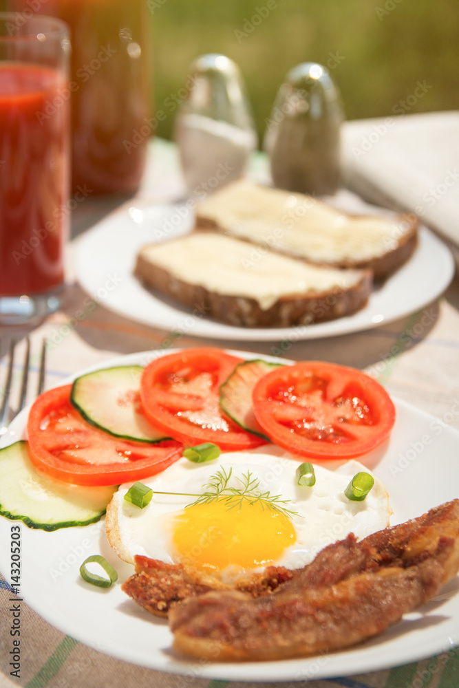 Morning breakfast: fried egg with ham, with sliced tomatoes and cucumbers, bread and butter, tomato juice in glass and bottle with newspaper on background. Cutlery, salt and pepper sunlight