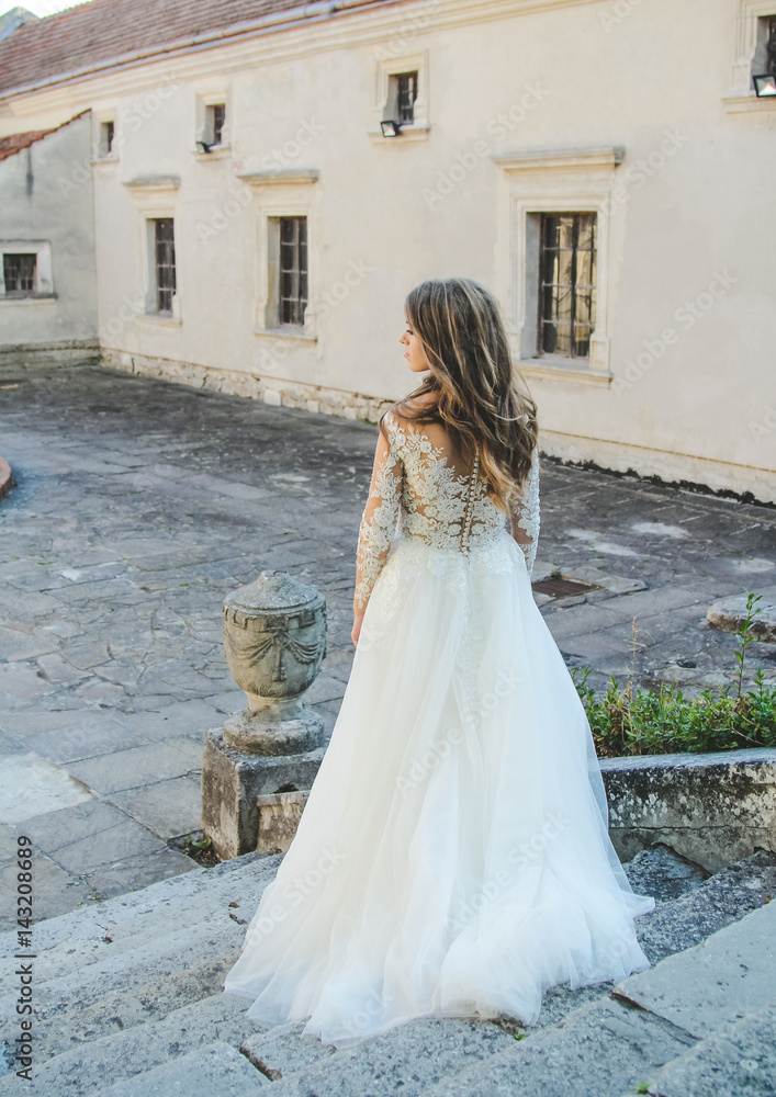 Back view of gorgeous bride standing in old stone castle. Beautiful, stylish girl in her sunny wedding day.