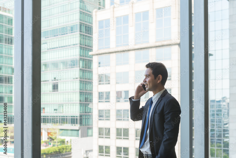 Mature and confident business executive looking looking out of large windows at a view of the city below, from the top floor of an office building, while talking on his mobile phone Mature and confident business executive looking looking out of large windows at a view of the city below, from the top floor of an office building, while talking on his mobile phone