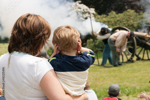 Woman holds her young son who blocks his ears as they watch canon firing at Civil War reenactment