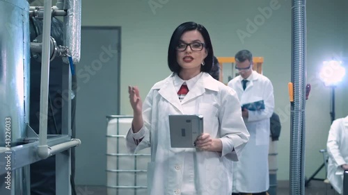 Female scientist in glasses and white lab coat holding tablet and walking through lab towards camera while talking and pointing to productional equipment photo