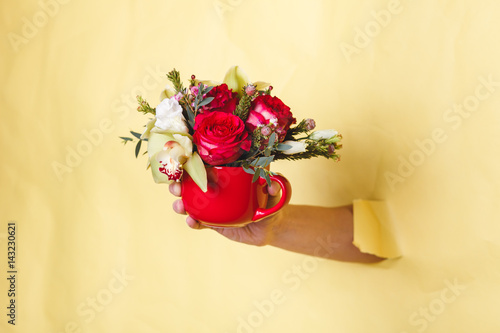 Fototapeta Naklejka Na Ścianę i Meble -  Hand holds small vase with flowers and rose breakes through a yellow background