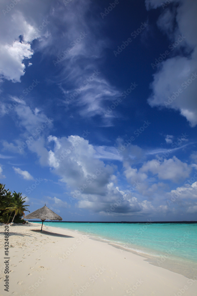 Fototapeta premium Straw umbrella on the white sandy beach on the coast of bright turquoise ocean and cloudy sky, Maldives