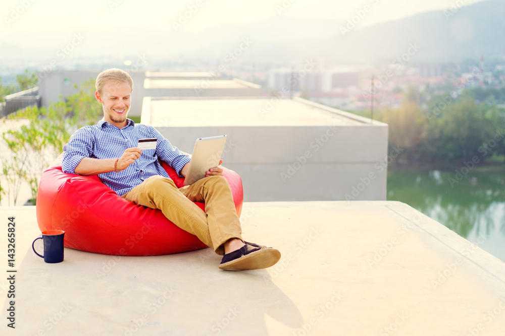 Shopping online. Internet banking. Handsome young man using tablet computer holding credit card while sitting on red beanbag on the roof top.