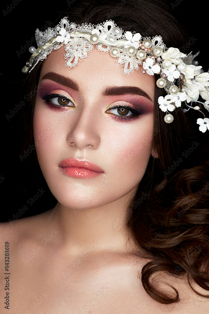 A young girl in the image of a bride with a floral wreath on her head and curls. A beautiful model with a bright makeup and perfect skin. Beauty of the face. Photo is taken in a studio.