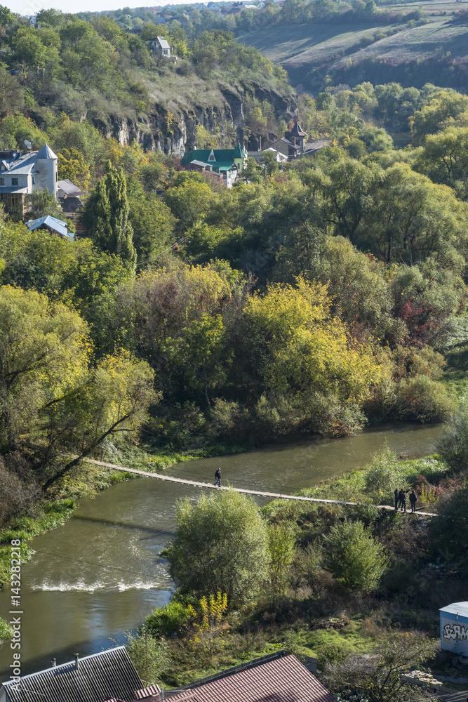Man walks across a bridge over the Smotrych River in the canyon of Kamianets-Podilskyi, Western Ukraine. Shot in autumn the trees are in full colour.