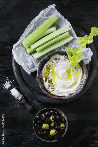 Fresh celery with yogurt and olive oil dip in ceramic bowl, served with sea salt and black, green olives on wood chopping board over black wooden burnt background. Top view with space. Healthy snack