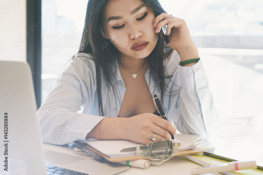 SMM specialist is making business calls by smartphone from the home office. Beautiful model look asian woman is sitting at the table with a laptop on it while talking by a cellphone. SMM specialist is making business calls by smartphone from the home office. Beautiful model look asian woman is sitting at the table with a laptop on it while talking by a cellphone.