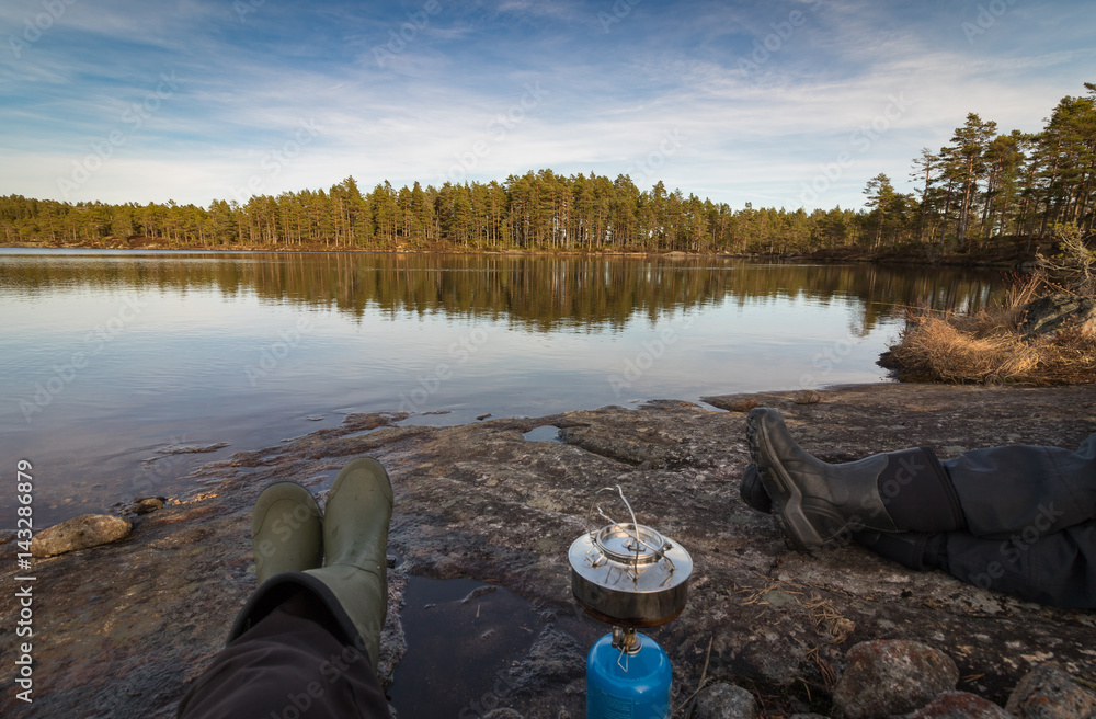 Obraz premium Two persons sitting at the edge of lake making coffe on a camp stove, pine forest in the wilderness of Norway