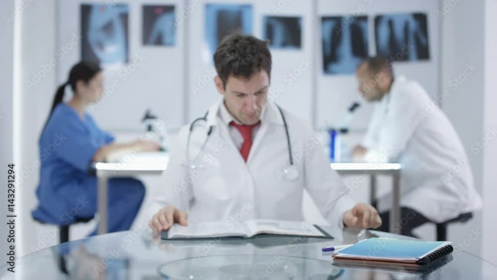 Portrait smiling doctor sitting at desk with colleagues working in background Portrait smiling doctor sitting at desk with colleagues working in background