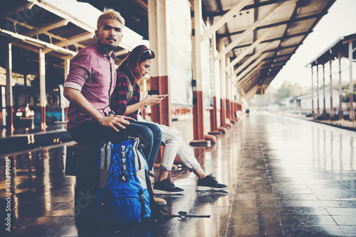 Young tourist couple with backpack sitting on bench at the train station. Two young tourist are waiting to get on the train and begin their journey. Travel concept.