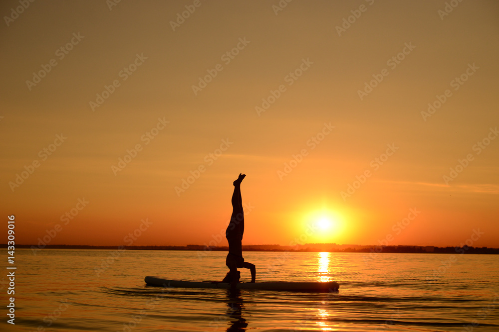 Beautiful young sporty woman trains upside down on the surf desk at sunset time