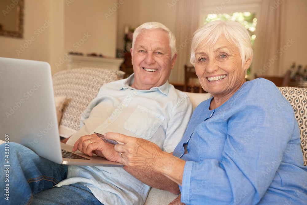Smiling seniors sitting at home shopping online with a laptop